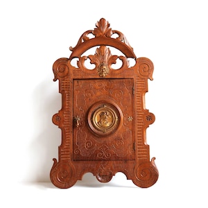 Könnte beinhalten: Ein geschnitzter Holzschrank mit einem dekorativen Rahmen und einer kleinen Tür. Die Tür hat ein rundes, verziertes Messingdesign in der Mitte.