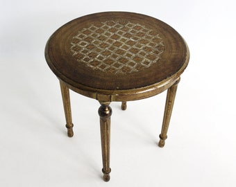 Puede incluir: Una mesa auxiliar redonda con una superficie de madera marrón oscuro que presenta un patrón geométrico de diamantes en color claro. La mesa tiene tres patas delgadas y cónicas con un acabado dorado desgastado y detalles tallados.
