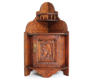 May include: A wooden corner cabinet with a carved door featuring a knight's helmet and intricate details. The cabinet has a small shelf and a brass knob.