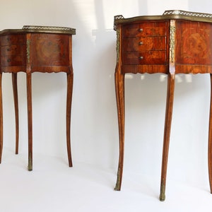 K&ouml;nnte beinhalten: Zwei antike Holz-Beistelltische mit jeweils drei Schubladen. Die Tische haben ein Marquetterie-Design und Messing-Akzente. Die Tische stehen auf einem wei&szlig;en Hintergrund.