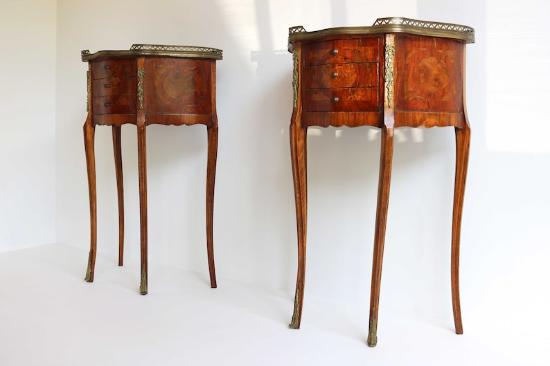 K&ouml;nnte beinhalten: Zwei antike Holz-Beistelltische mit jeweils drei Schubladen. Die Tische haben ein Marquetterie-Design und Messing-Akzente. Die Tische stehen auf einem wei&szlig;en Hintergrund.