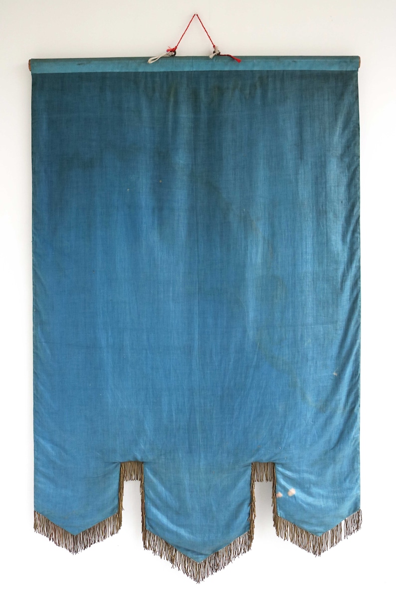 May include: A blue fabric banner with a fringed bottom edge. The banner is hanging from a wooden dowel rod with a red string.
