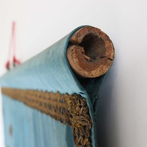 May include: A blue fabric scroll with gold trim, rolled up and secured with a wooden dowel. The scroll is hanging from a red string.