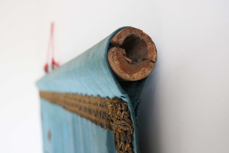 May include: A blue fabric scroll with gold trim, rolled up and secured with a wooden dowel. The scroll is hanging from a red string.