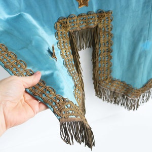 May include: A teal blue fabric with gold trim and fringe. The fabric has a decorative pattern and is likely part of a costume or ceremonial garment.
