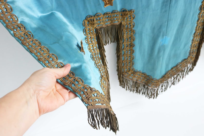 May include: A teal blue fabric with gold trim and fringe. The fabric has a decorative pattern and is likely part of a costume or ceremonial garment.