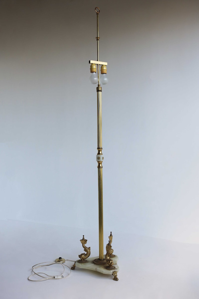 May include: A vintage brass floor lamp with a marble base and dolphin accents. The lamp features a slender, adjustable pole with two light bulbs. The base has a triangular shape with decorative feet. The lamp is approximately 60 inches tall.