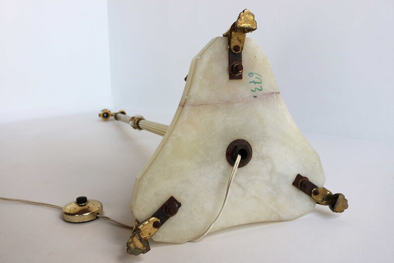 May include: A vintage table lamp with a triangular, off-white marble base and a slender, fluted column. The lamp features gold-toned metal accents and a foot switch. The lamp's base has a visible crack and the number "673" is written on the side.