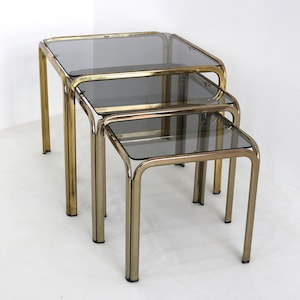 May include: Set of three nesting tables with a gold-toned metal frame and smoked glass tops. The tables are square and have a minimalist design, perfect for a living room or a modern space. The largest table is approximately 20 inches wide.