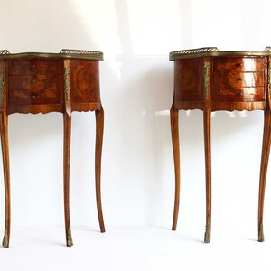 K&ouml;nnte beinhalten: Zwei antike Holz-Beistelltische mit jeweils drei Schubladen. Die Tische haben ein Marquetterie-Design und Messing-Akzente. Die Tische stehen auf einem wei&szlig;en Hintergrund.