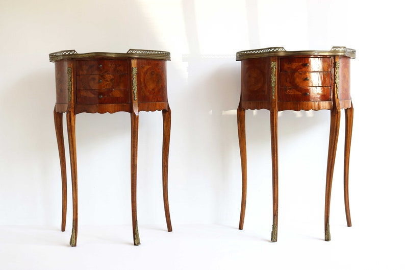 K&ouml;nnte beinhalten: Zwei antike Holz-Beistelltische mit jeweils drei Schubladen. Die Tische haben ein Marquetterie-Design und Messing-Akzente. Die Tische stehen auf einem wei&szlig;en Hintergrund.