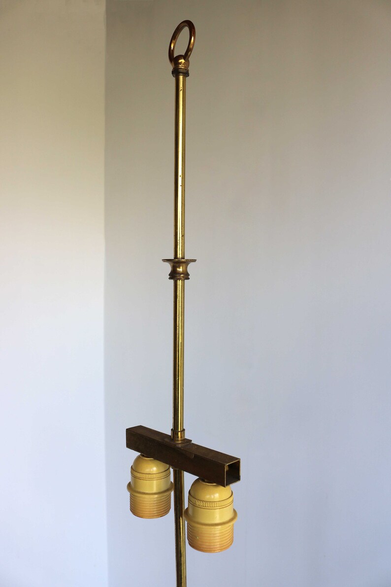 May include: A vintage brass floor lamp with a ring at the top and two light bulb sockets. The lamp has a long, slender pole and a rectangular bar holding the sockets. The sockets are a light yellow color.