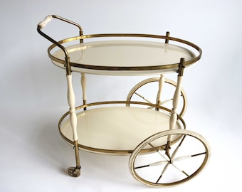 May include: A two-tiered serving cart with a brass frame and white lacquered trays. The cart has four wheels, two of which are visible in the image. The cart is on a white background.