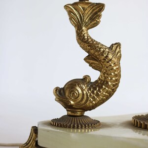 May include: A gold-colored, ornate lamp base featuring a stylized fish design. The lamp base has a rectangular, off-white surface and a decorative foot. The fish is detailed with scales and fins.