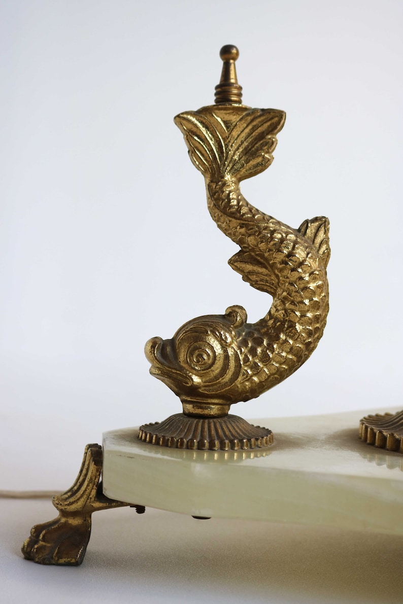 May include: A gold-colored, ornate lamp base featuring a stylized fish design. The lamp base has a rectangular, off-white surface and a decorative foot. The fish is detailed with scales and fins.