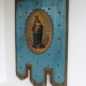 May include: A blue fabric banner with gold trim and fringe. The banner features a painting of the Virgin Mary holding a baby Jesus. The painting is surrounded by a gold frame and stars.
