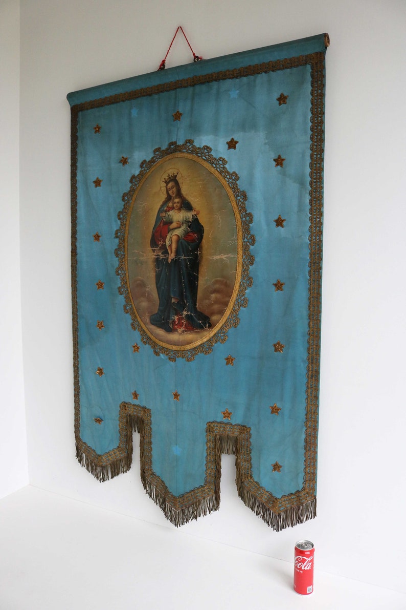 May include: A blue fabric banner with gold trim and fringe. The banner features a painting of the Virgin Mary holding a baby Jesus. The painting is surrounded by a gold frame and stars.