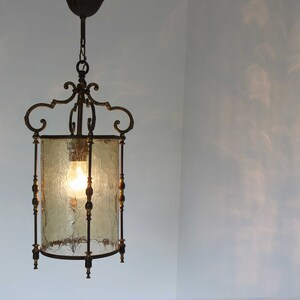 Pode incluir: Uma lumin&aacute;ria pendente de lanterna de lat&atilde;o vintage com um abajur de vidro fosco. A lanterna tem um design decorativo e ornamentado com uma corrente pendurada no teto.