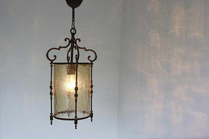 Pode incluir: Uma lumin&aacute;ria pendente de lanterna de lat&atilde;o vintage com um abajur de vidro fosco. A lanterna tem um design decorativo e ornamentado com uma corrente pendurada no teto.