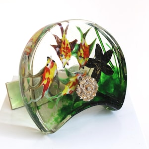 May include: A clear, curved glass paperweight with a painted underwater scene, including colourful fish and green plants. A gold-coloured, decorative embellishment is in the centre. The paperweight is on a white surface.