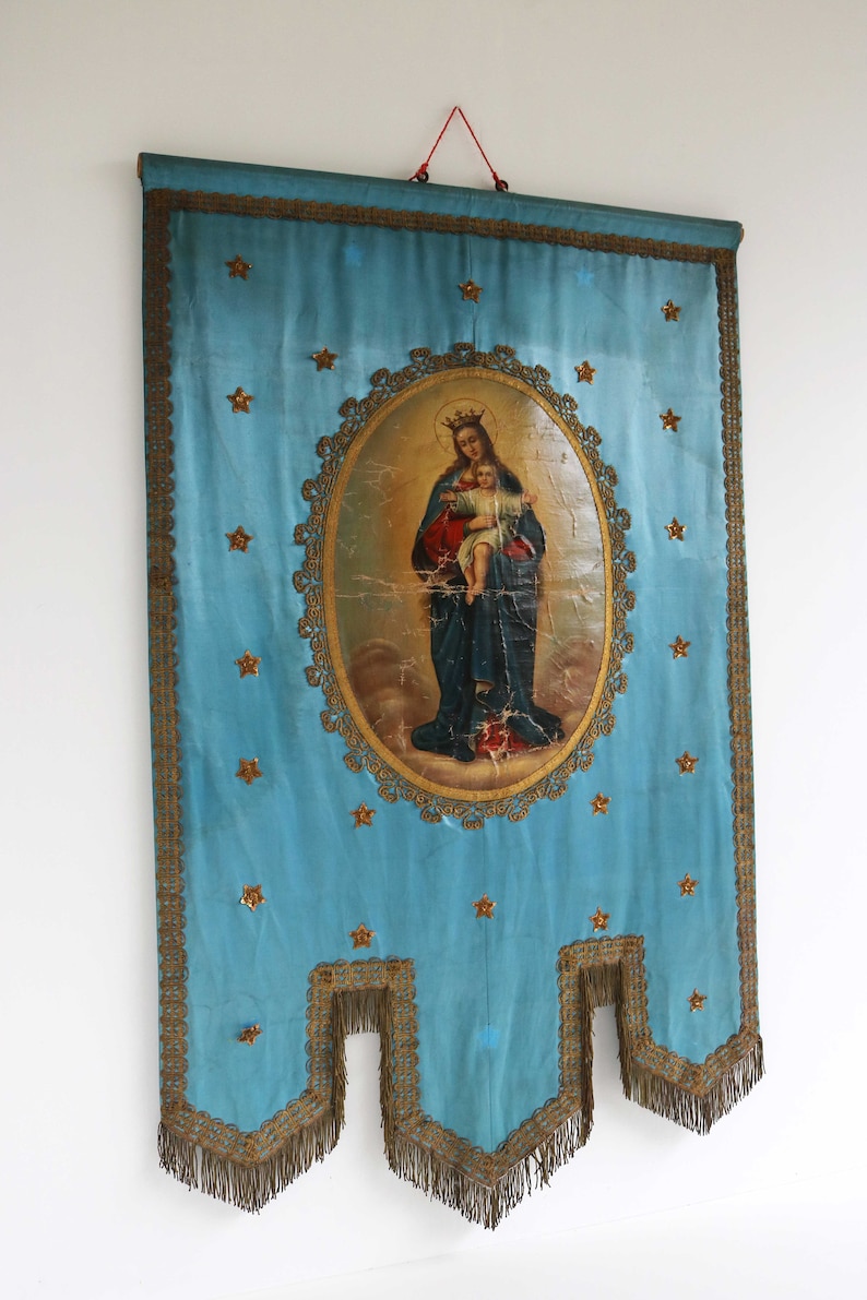 May include: A blue fabric banner with gold trim and a central image of the Virgin Mary holding a baby Jesus. The image is surrounded by a gold frame and the banner is decorated with gold stars.
