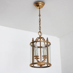 May include: A gold-toned, cylindrical pendant light fitting with clear glass panels. The fixture features two faux candle lights and is suspended from the ceiling by a chain. The design is ornate and vintage.