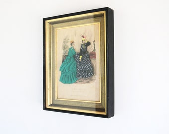 May include: Framed vintage fashion print featuring two women in period dresses. The print is in a black frame with a gold inner border. One woman wears a green dress, the other a black dress with white polka dots. The print is on a beige background.