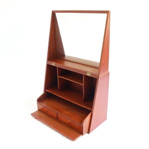 May include: A mid-century wooden vanity unit with a mirror. The unit has a rectangular shape with a slanted base, two drawers, and open shelving. The mirror is framed by a tall, triangular structure.