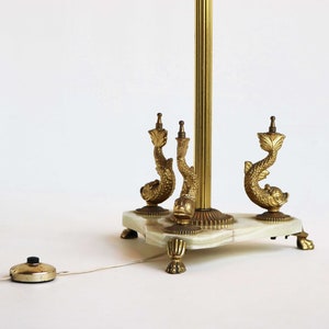 May include: A vintage brass floor lamp with a white marble base. The lamp features three decorative dolphin figures and a foot switch. The lamp's brass pole is fluted, and the base has ornate feet.