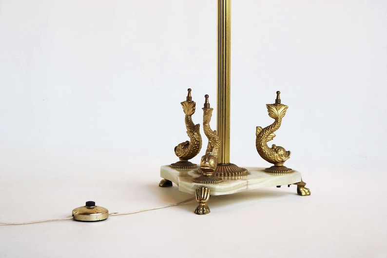 May include: A vintage brass floor lamp with a white marble base. The lamp features three decorative dolphin figures and a foot switch. The lamp's brass pole is fluted, and the base has ornate feet.