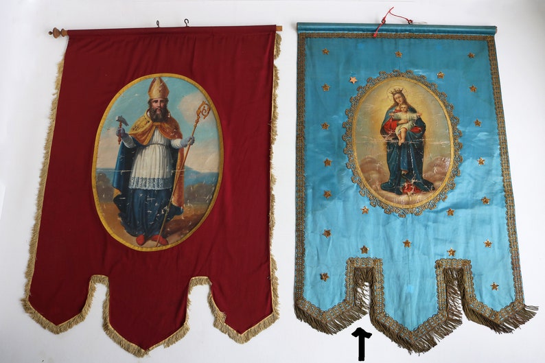 May include: Two vintage religious banners with gold trim and fringe. The banner on the left is red with a painting of a man in a bishop's robe holding a staff and a hammer. The banner on the right is blue with a painting of a woman holding a baby.