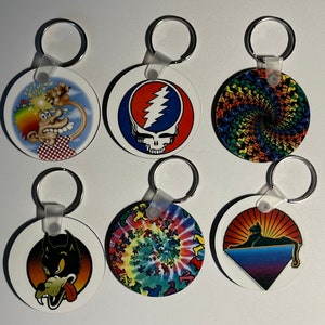 May include: Six round keychains with different colourful designs. The keychains feature a cartoon character with a rainbow on its head, a skull with a lightning bolt, a swirling rainbow pattern, a tie-dye pattern, a wolf with its tongue sticking out, and a black cat with a sunset background.