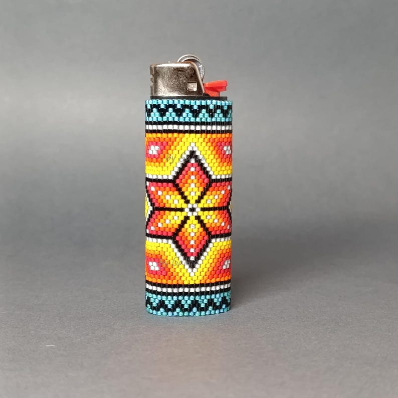 May include: A beaded lighter cover with a colorful geometric pattern featuring a large orange flower in the center. The cover is made of small, colorful beads and is designed to fit a standard-sized lighter.