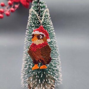 May include: A beaded Christmas ornament shaped like an owl wearing a red Santa hat. The owl is brown and orange with white eyes and a white beak. The ornament is hanging from a silver chain on a small artificial Christmas tree.