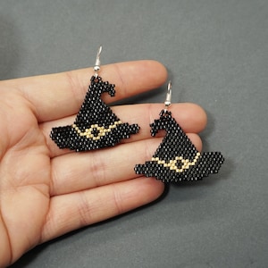 May include: A pair of black witch hat earrings with gold accents. The earrings are made of beads and have a silver hook.