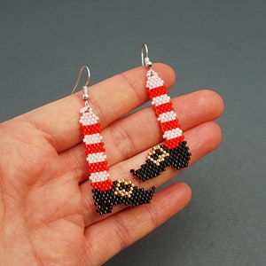 Op de afbeelding: Een paar grillige kralen oorbellen in de vorm van kerstmanbenen. De oorbellen hebben rood-wit gestreepte benen met zwarte laarzen en gouden details. De oorbellen hangen aan zilverkleurige haken.