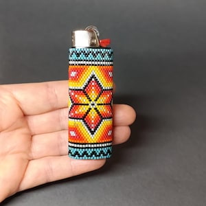 May include: A beaded lighter cover with a colorful geometric pattern featuring a large orange flower in the center. The cover is made of small, colorful beads and has a blue, black, and white border.