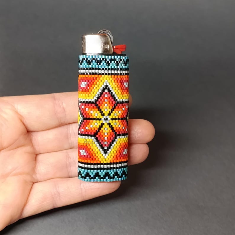 May include: A beaded lighter cover with a colorful geometric pattern featuring a large orange flower in the center. The cover is made of small, colorful beads and has a blue, black, and white border.