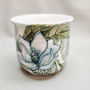 May include: A white ceramic cup with a hand-painted design of a blue and white magnolia flower and green leaves. The cup has a slightly flared rim and a smooth, glossy finish. The floral design wraps around the cup.