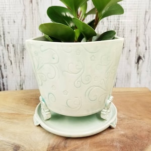 Peut inclure: Un pot de plante en céramique vert clair avec une soucoupe assortie. Le pot présente un motif floral en relief. Le pot est posé sur une surface en bois et contient une plante verte.