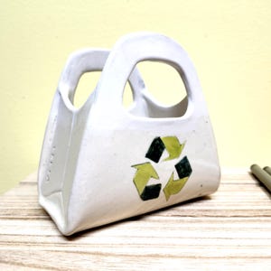 May include: White ceramic bag-shaped container with a green and black recycling symbol. The container has two handles. It is on a wooden surface. The background is a light yellow colour.