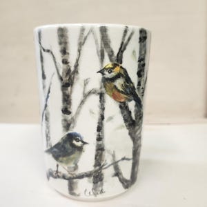 May include: A white ceramic tumbler featuring a hand-painted scene of two small birds perched on bare tree branches. One bird has yellow and brown plumage, while the other is blue and grey. The design includes black birch tree trunks and a signature.
