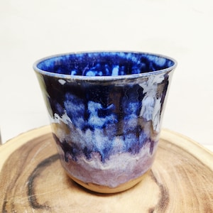 May include: A ceramic cup featuring a reactive glaze with a deep blue rim that transitions into lighter shades of blue and purple, ending in a natural tan base. The surface has a glossy finish with visible drips and blended color gradients.