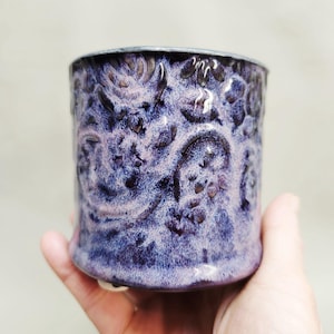 May include: A handmade ceramic mug with a textured, embossed floral pattern. The mug has a deep purple glaze with lighter purple highlights, and a dark rim. The mug is held in a hand, showing its size and shape.