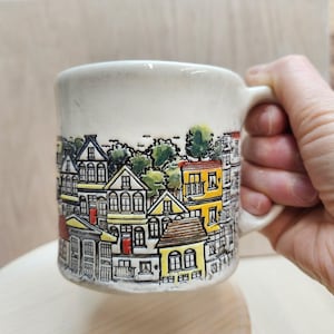 May include: Cream-colored ceramic mug featuring a detailed illustration of a townscape with houses in yellow, white, and red. The mug has a curved handle and a slightly textured surface, perfect for coffee or tea.
