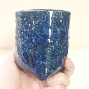 May include: A cylindrical ceramic mug featuring a deep blue glaze with a textured surface. The exterior is decorated with embossed celestial motifs, including multiple stars and crescent moons scattered across the dark, glossy finish.