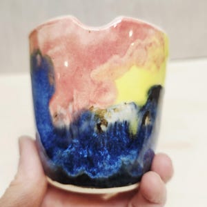 May include: A ceramic cup featuring a glossy, multi-colored glaze with blended sections of deep blue, soft pink, and bright yellow. The rim has a small, distinct indentation, and the surface displays a smooth, reflective finish throughout.
