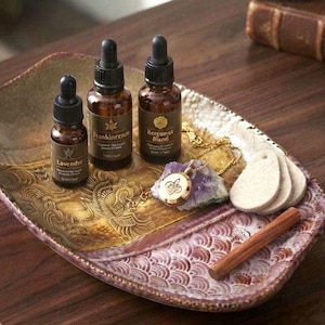 May include: Three amber glass dropper bottles labelled Lavender, Frankincense, and Bergamot Blend, a gold-tone pendant necklace resting on a purple amethyst crystal, three white felt discs, and a wooden stick on a decorative ceramic tray.