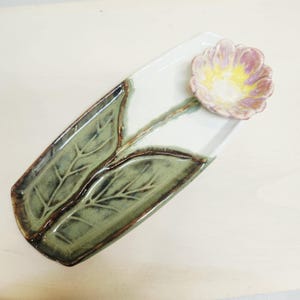 May include: A rectangular ceramic tray with a white base featuring two sculpted green leaves with dark brown edges. A three-dimensional flower with pink and yellow petals is attached to the top right corner, connected by a thin green stem.