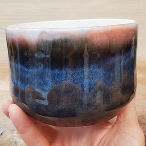 May include: A handcrafted ceramic bowl with a distinctive glaze. The bowl displays a colour gradient of white, red, blue, and black. The interior is white, and the exterior has a glossy finish. This bowl is suitable for serving food or as a decorative item.